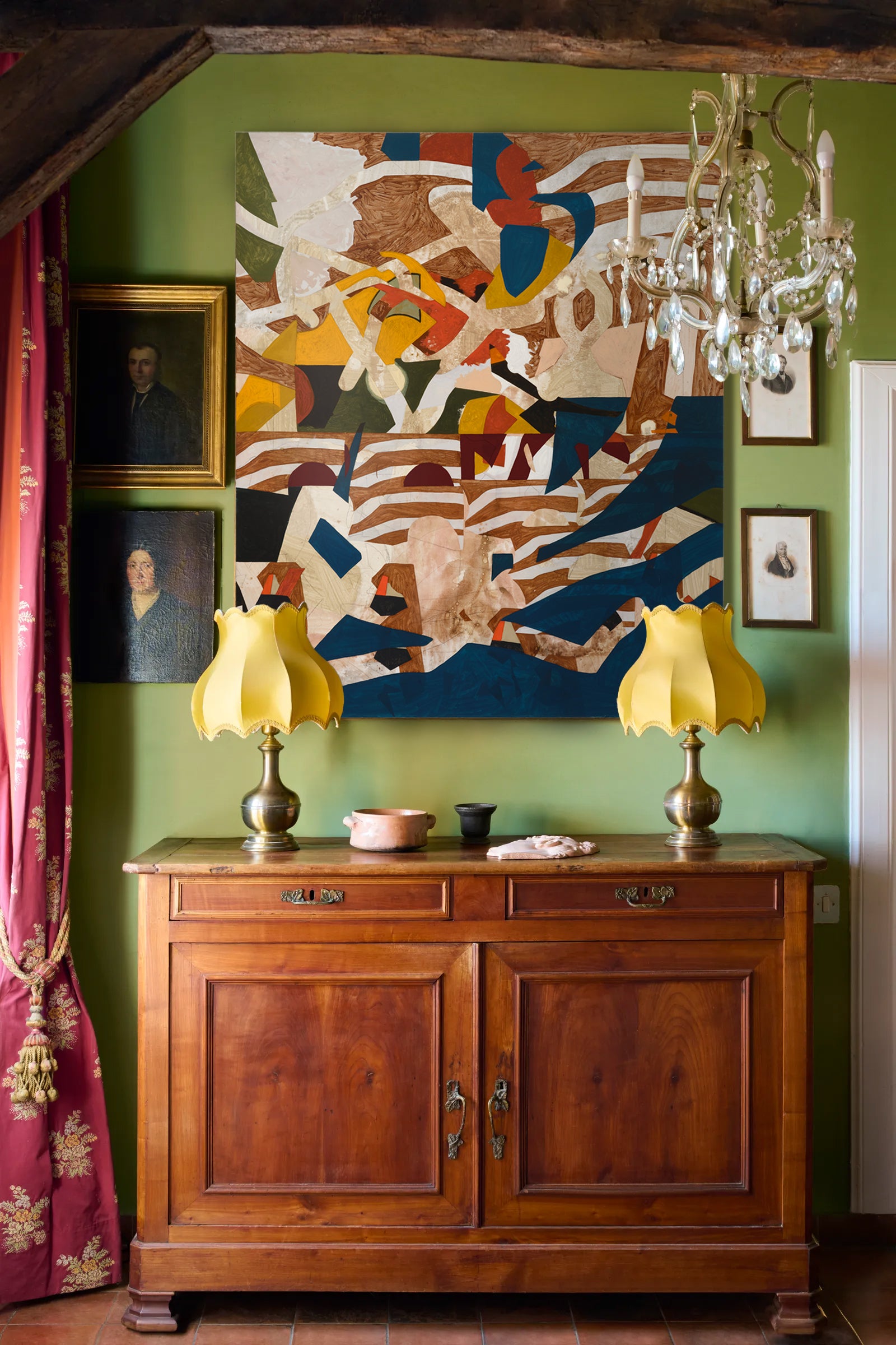 Wooden sideboard with two yellow lamps against a green wall with abstract art and framed pictures.